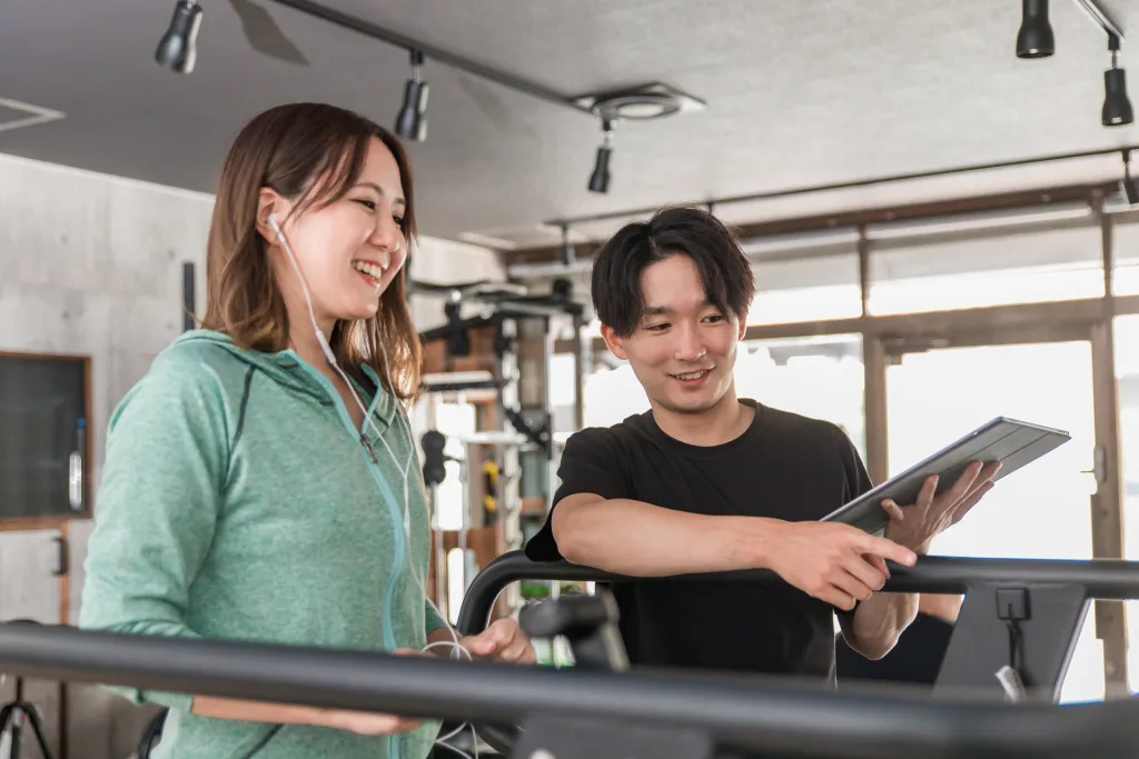 Two people at a gym smiling and chatting; a woman in a green hoodie with earbuds and a man in a black shirt shows a tablet and points.